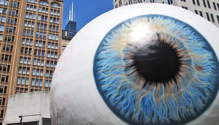 A giant statue of an eyeball sits in front of skyscrapers and a blue sky
