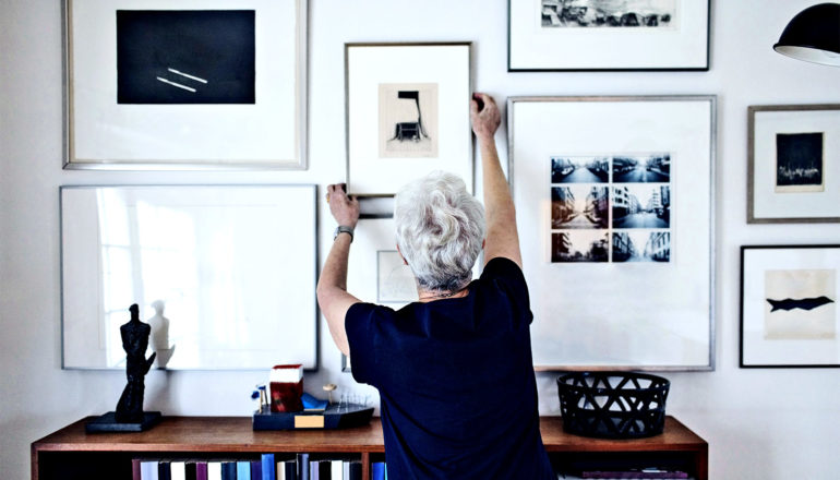An older woman hangs a framed photograph on a wall covered in other framed art