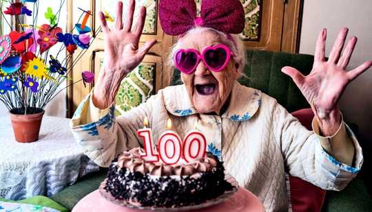 A woman looks excited with pink glasses and a bow on, looking at a cake with 