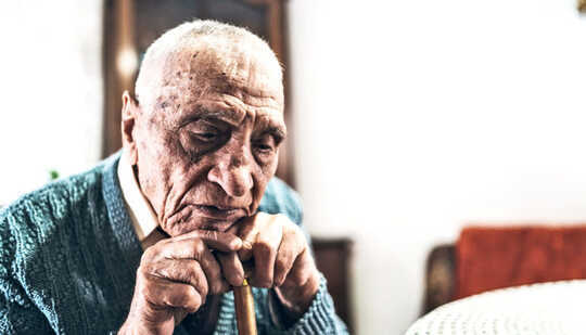 An older man rests his chin on his cane while sitting down at a table
