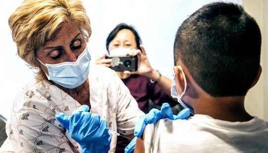 A nurse gives a child the COVID-19 vaccine as his mother takes a photo with her phone