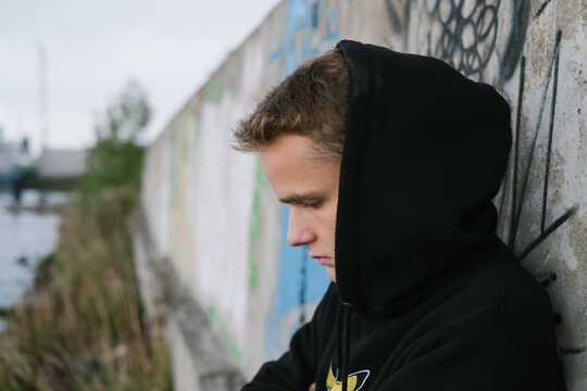 Teenage boy in a hoodie stands against a wall, looking down
